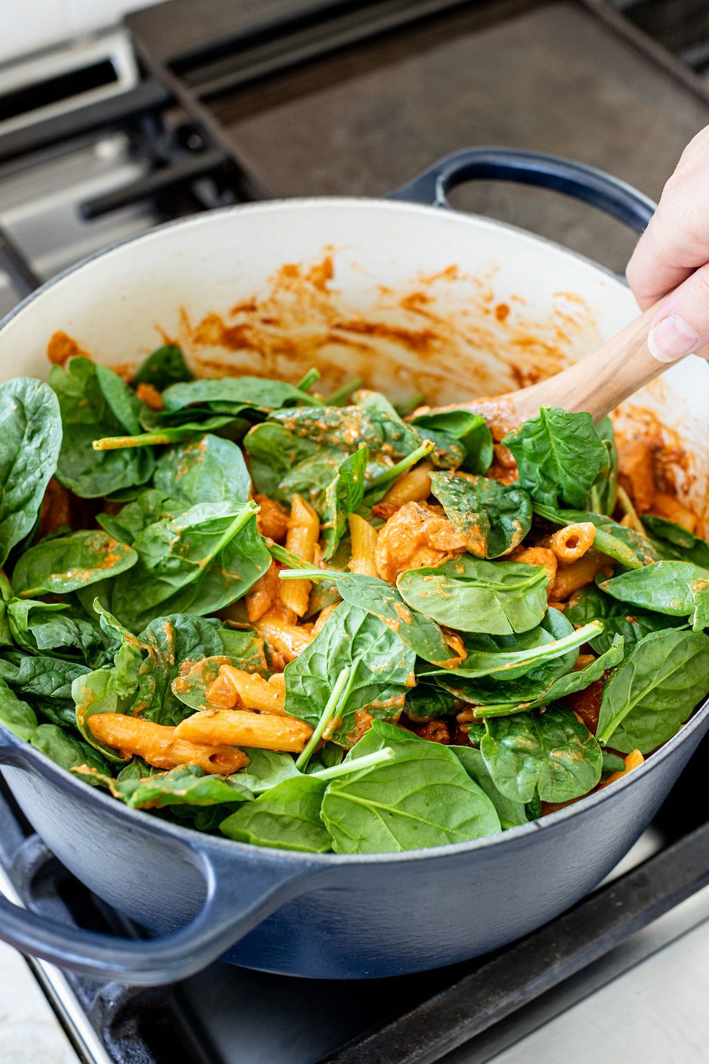 A hand stirring fresh baby spinach into creamy orange pasta.