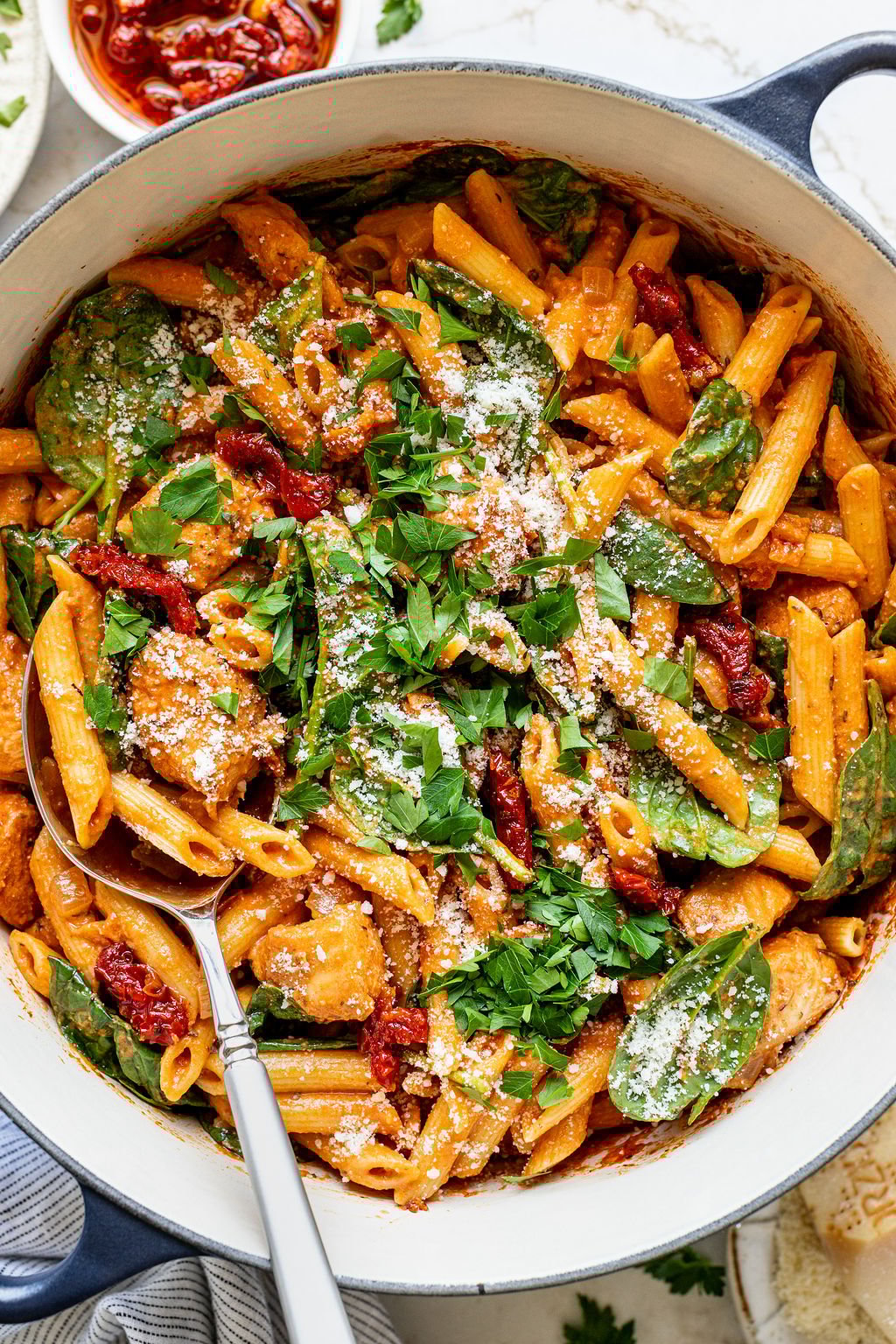 A spoon in a pot of creamy orange pasta with baby spinach, sun dried tomatoes, and chunky spiced chicken.