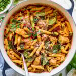 A spoon in a pot of with creamy tomato chicken pasta next to bowls of sundried tomatoes and basil.