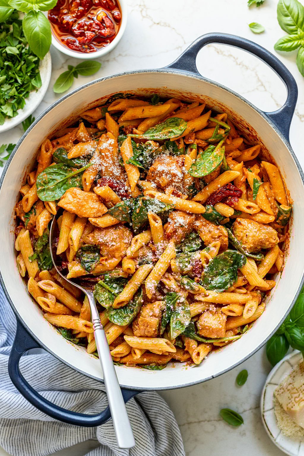 A spoon in a pot of with creamy tomato chicken pasta next to bowls of sundried tomatoes and basil.