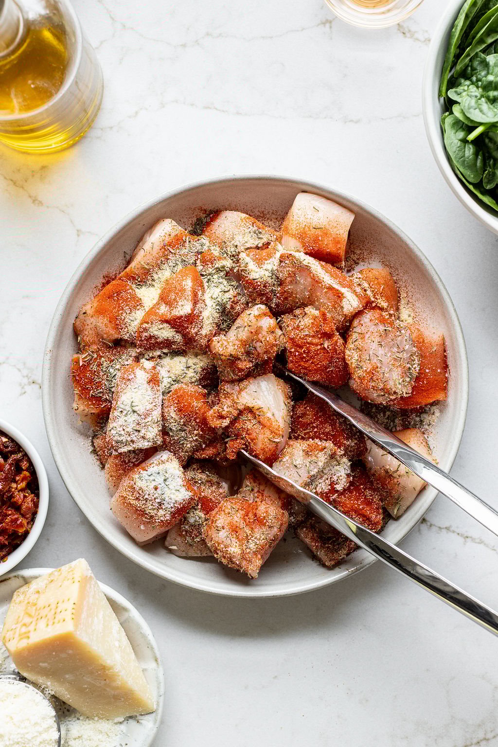 Tongs tossing chunky chicken breast in spices.