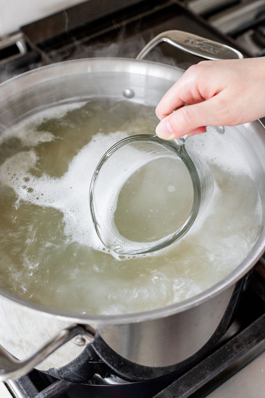 A hand scooping pasta water from a pot with a glass measuring cup.