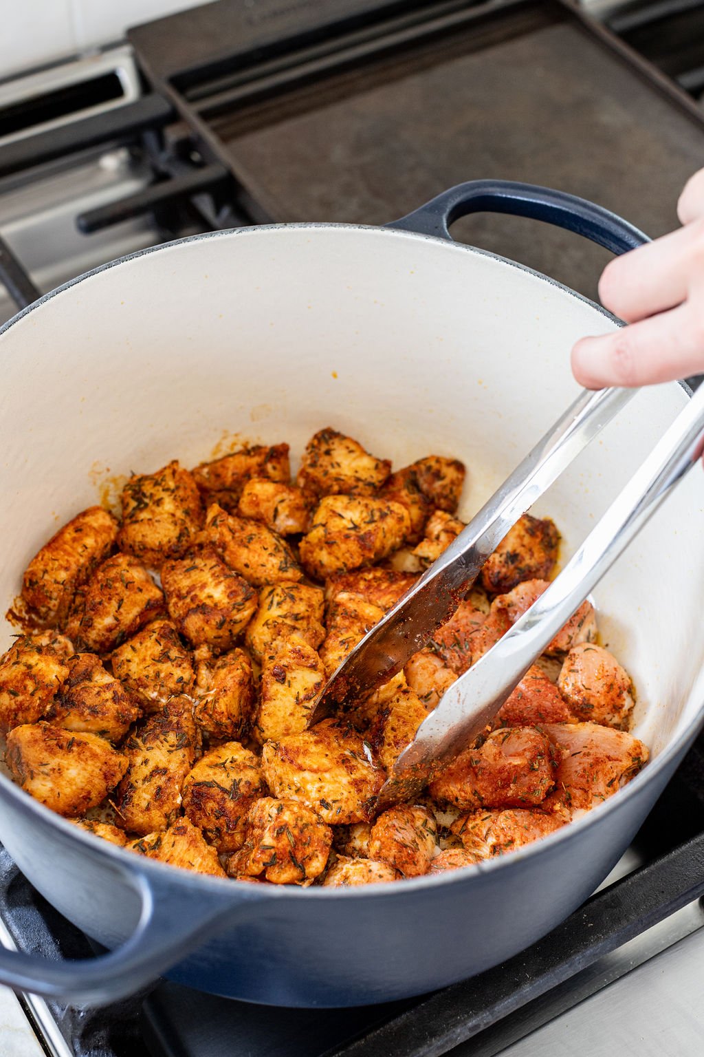 A hand turning cooked chicken pieces in a large pot with tongs.