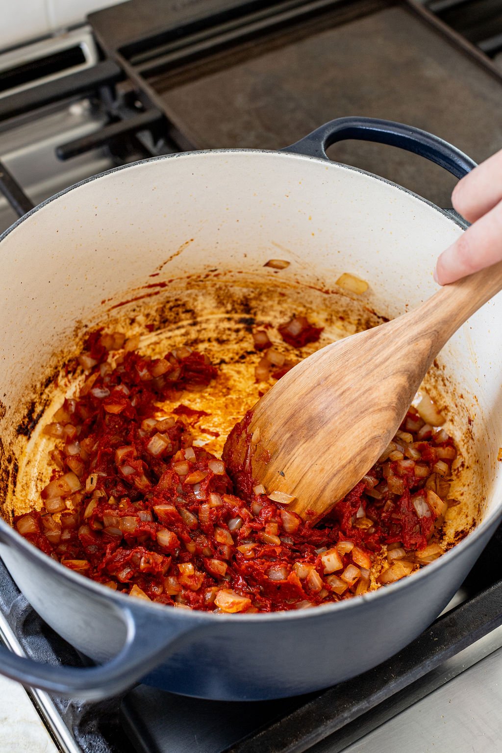 A hand using a wooden spoon to stir tomato paste into cooked diced onions in a pot.