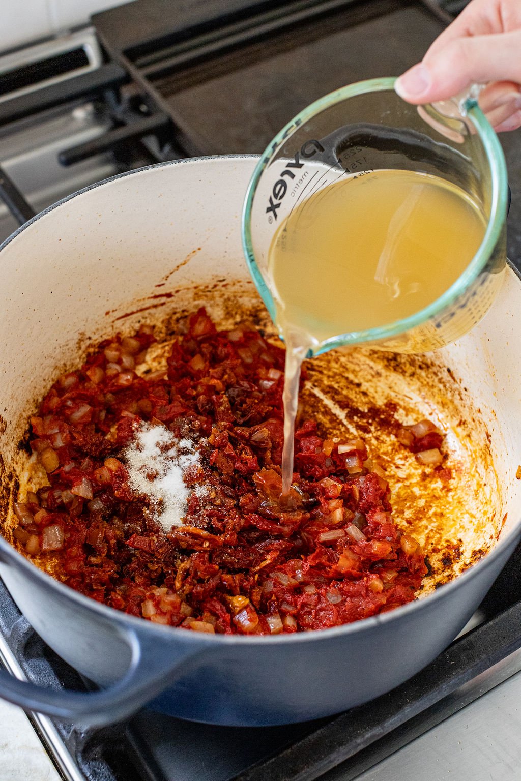 Chicken broth being poured into a pot with tomato sauce, spices, sugar, and diced onions.