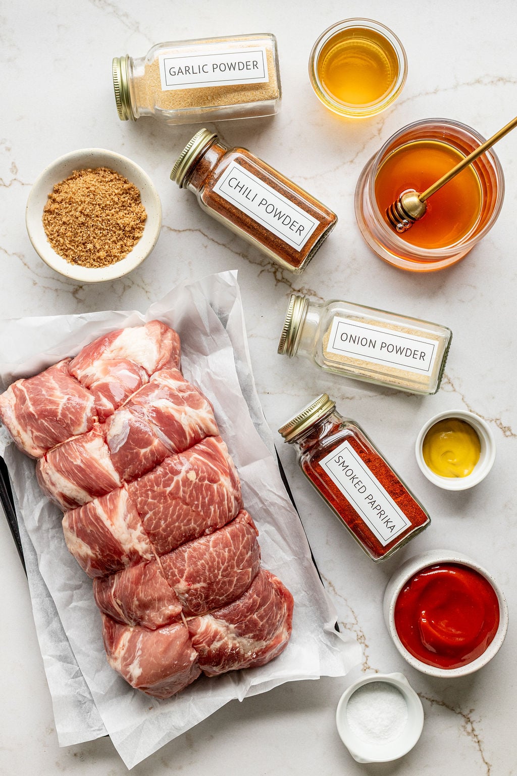 Raw pork next to bowls and jars of honey, mustard, salt, and spices on a table.