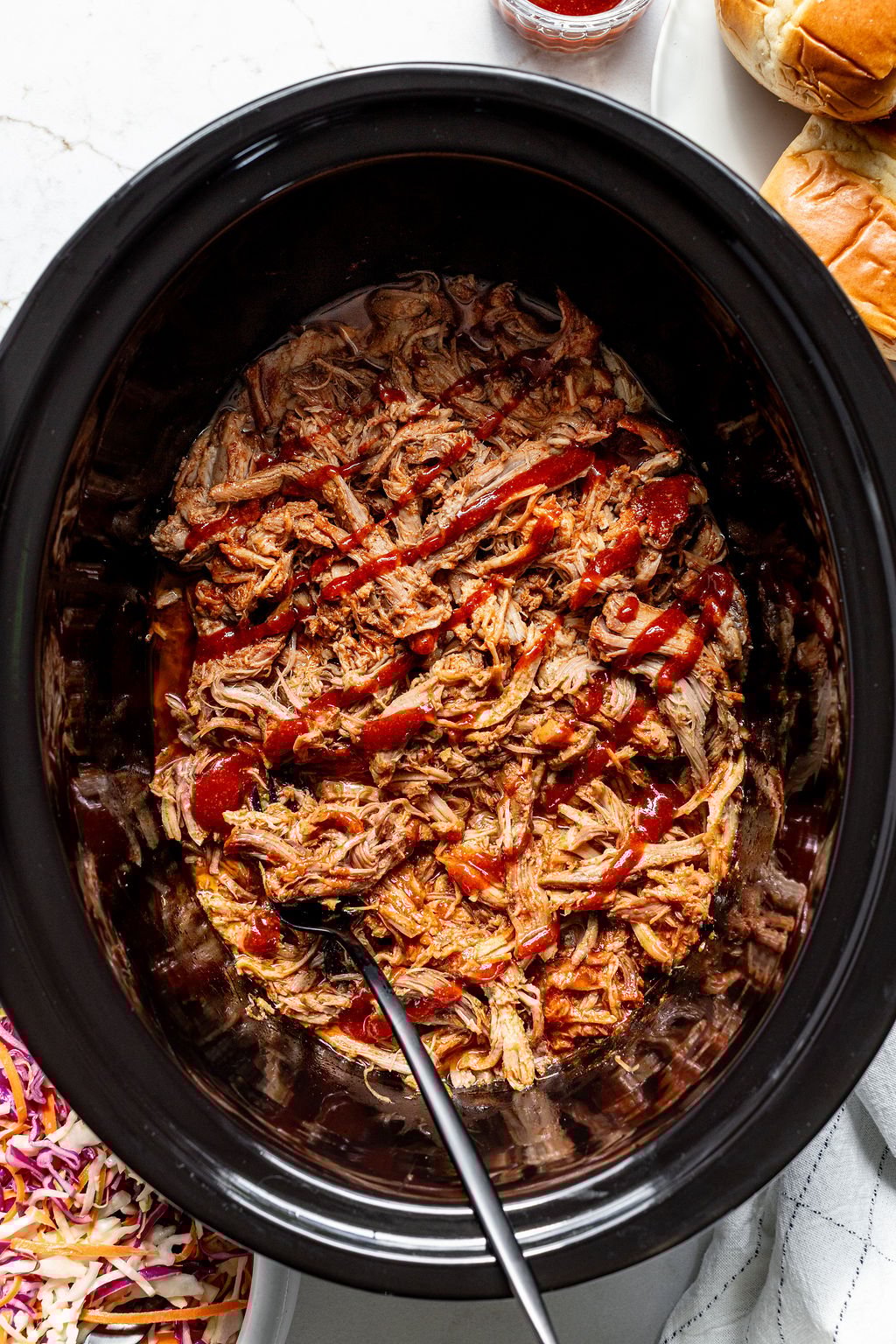 A slow cooker filled with barbecue shredded pork next to buns and coleslaw.