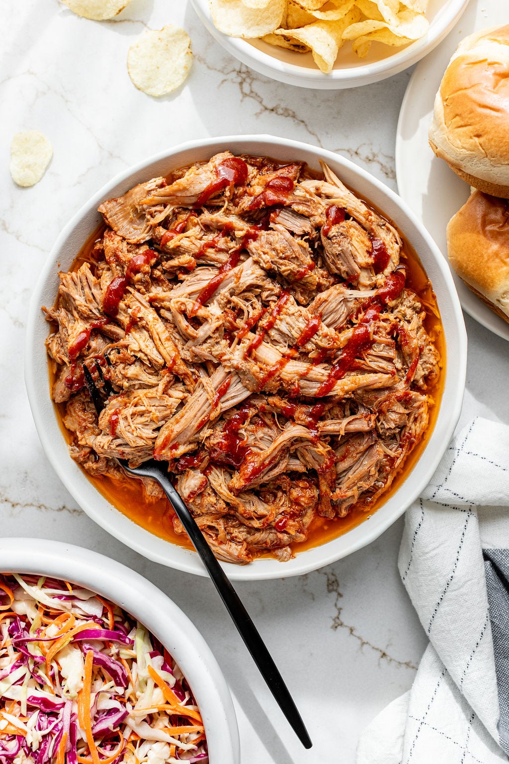 Juicy shredded pork in a shallow bowl next to burger buns and coleslaw.