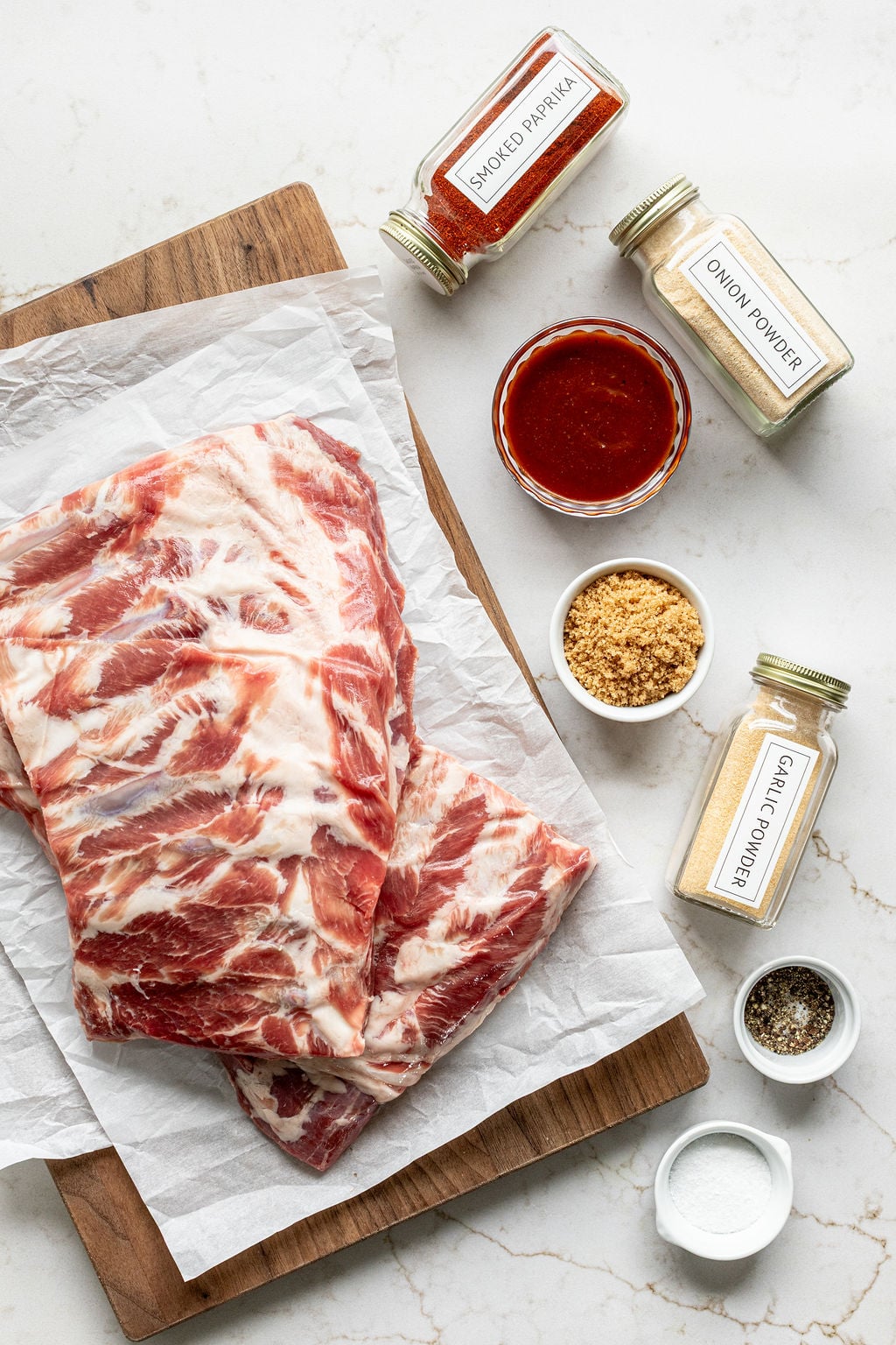 Raw pork ribs next to jars and bowls of spices on a table.
