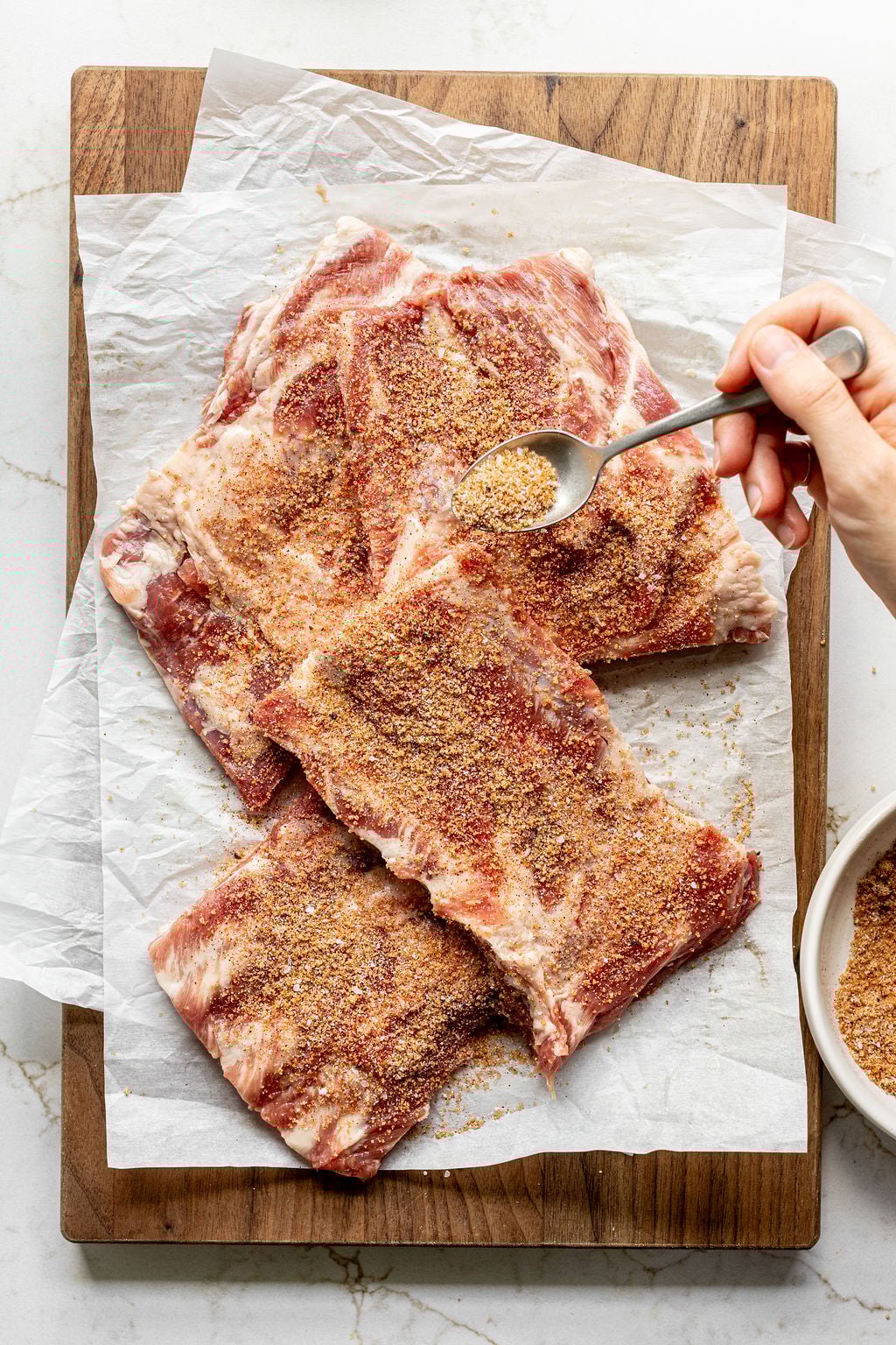 A hand sprinkling a brown sugar spice rub onto pork ribs.