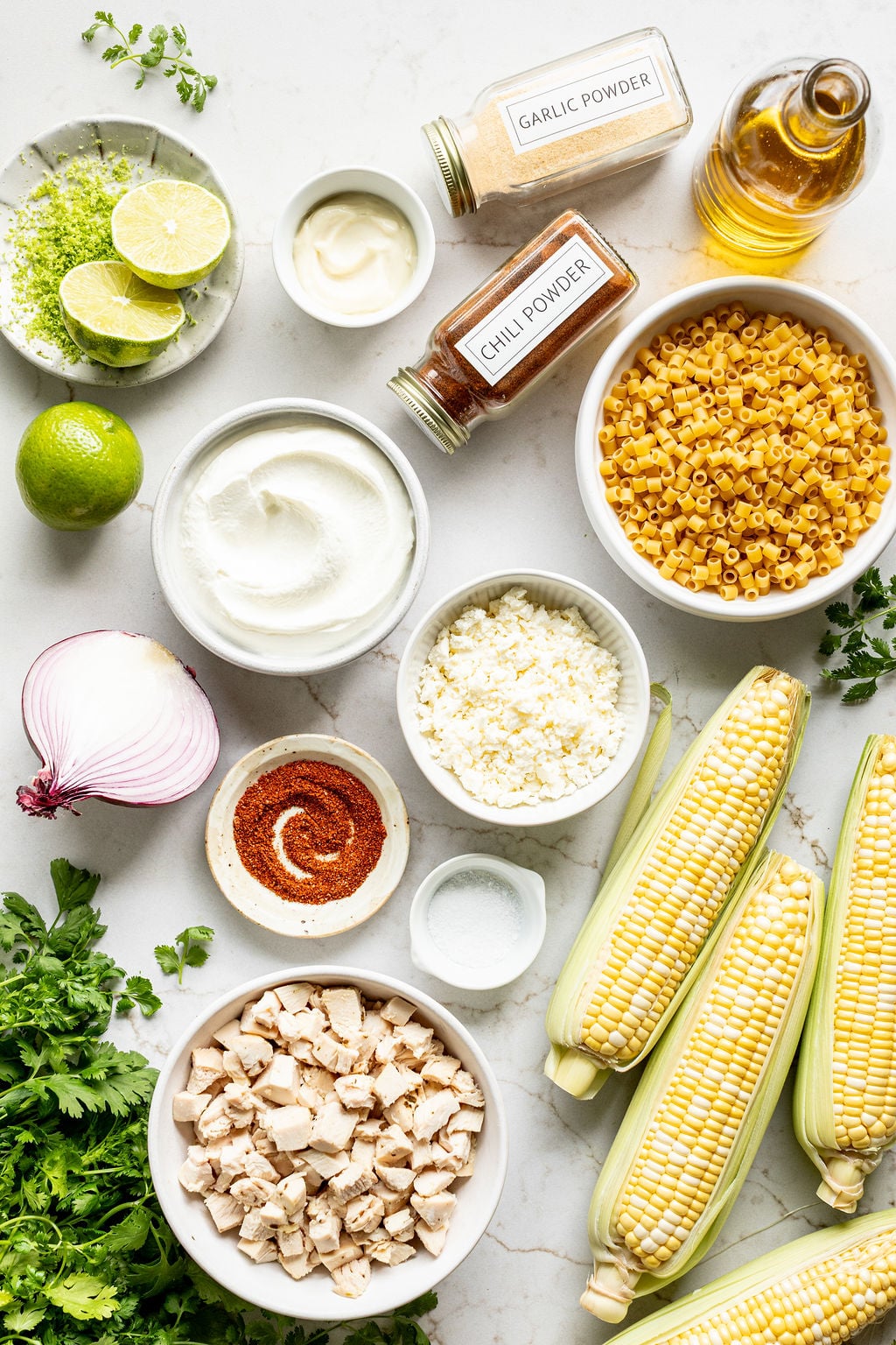 Corn on the cob next to bowls of diced chicken, pasta, lime zest, cotija, Greek yogurt, and spices.