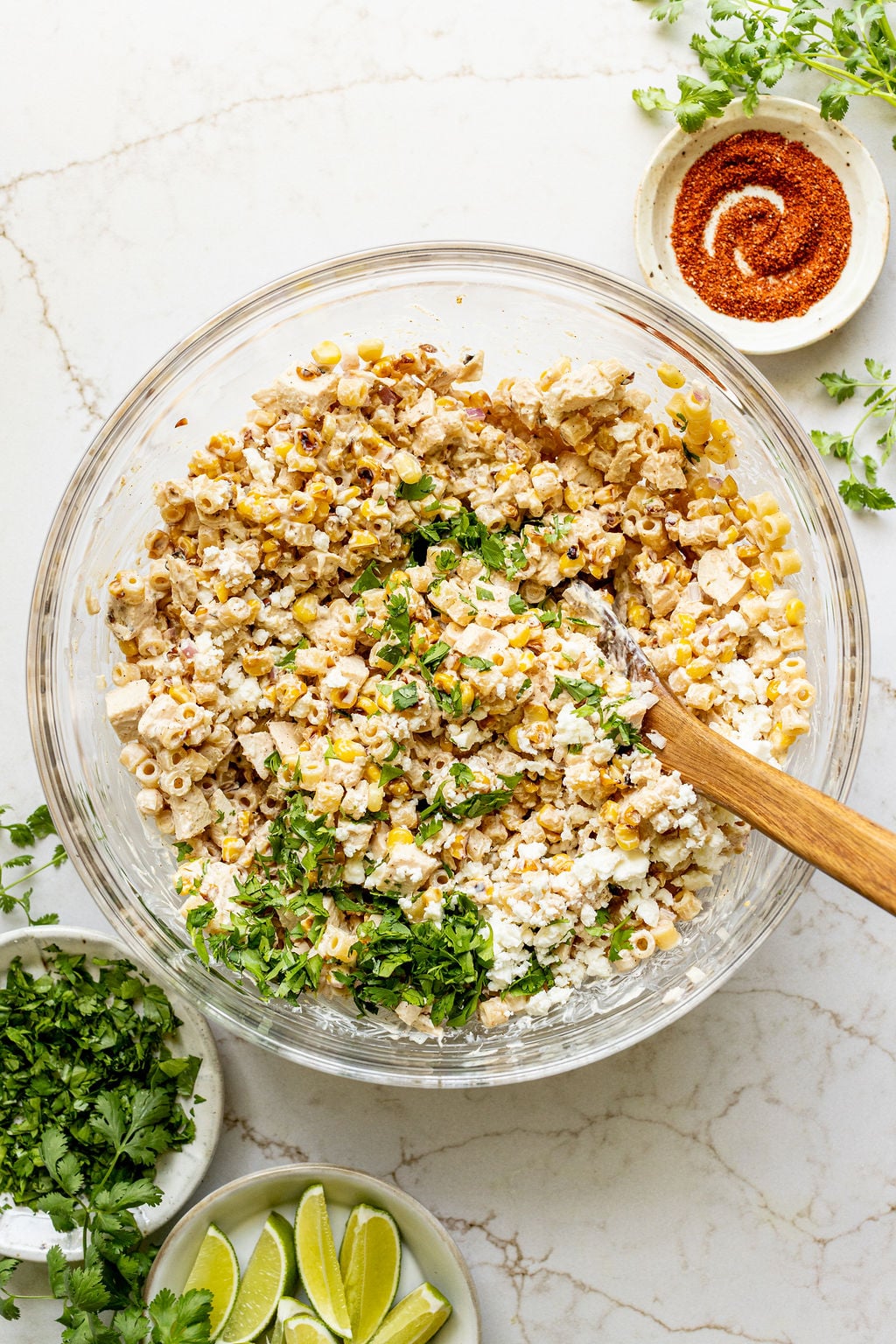 A spoon stirring cotija cheese and chopped cilantro into a bowl of corn pasta salad.