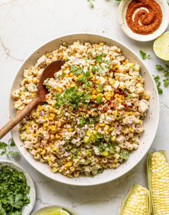 Corn on the cob next to a large bowl filled with pasta, charred corn, diced chicken, cotija cheese, and a creamy lime spice yogurt mixture.