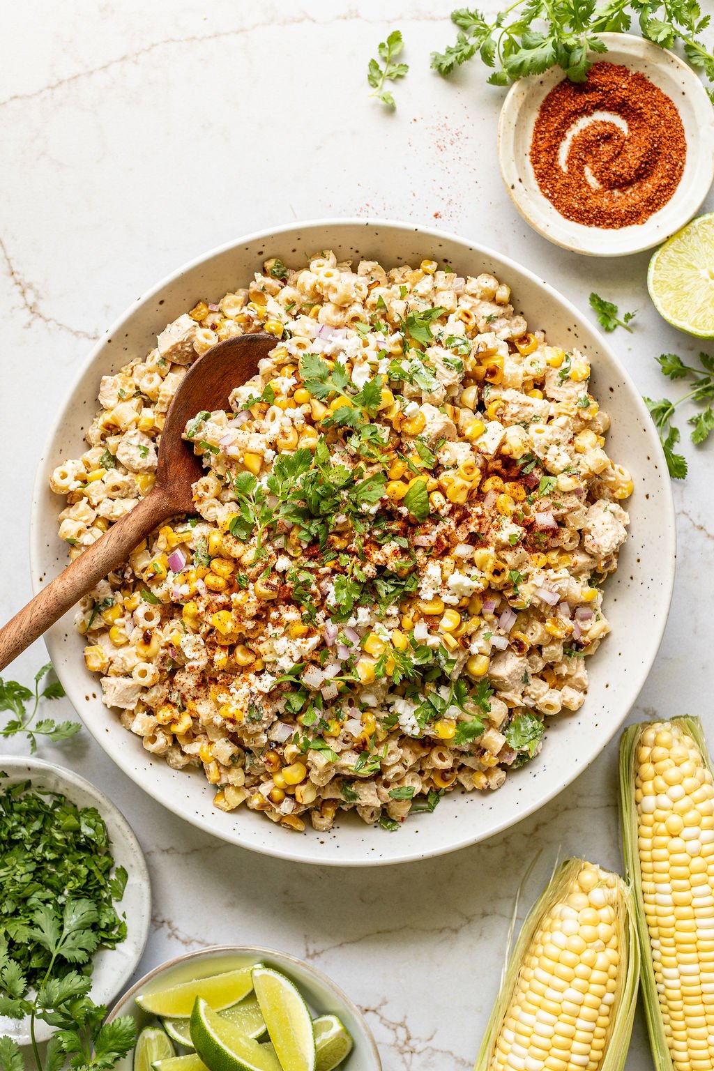 Corn on the cob next to a large bowl filled with pasta, charred corn, diced chicken, cotija cheese, and a creamy lime spice yogurt mixture.