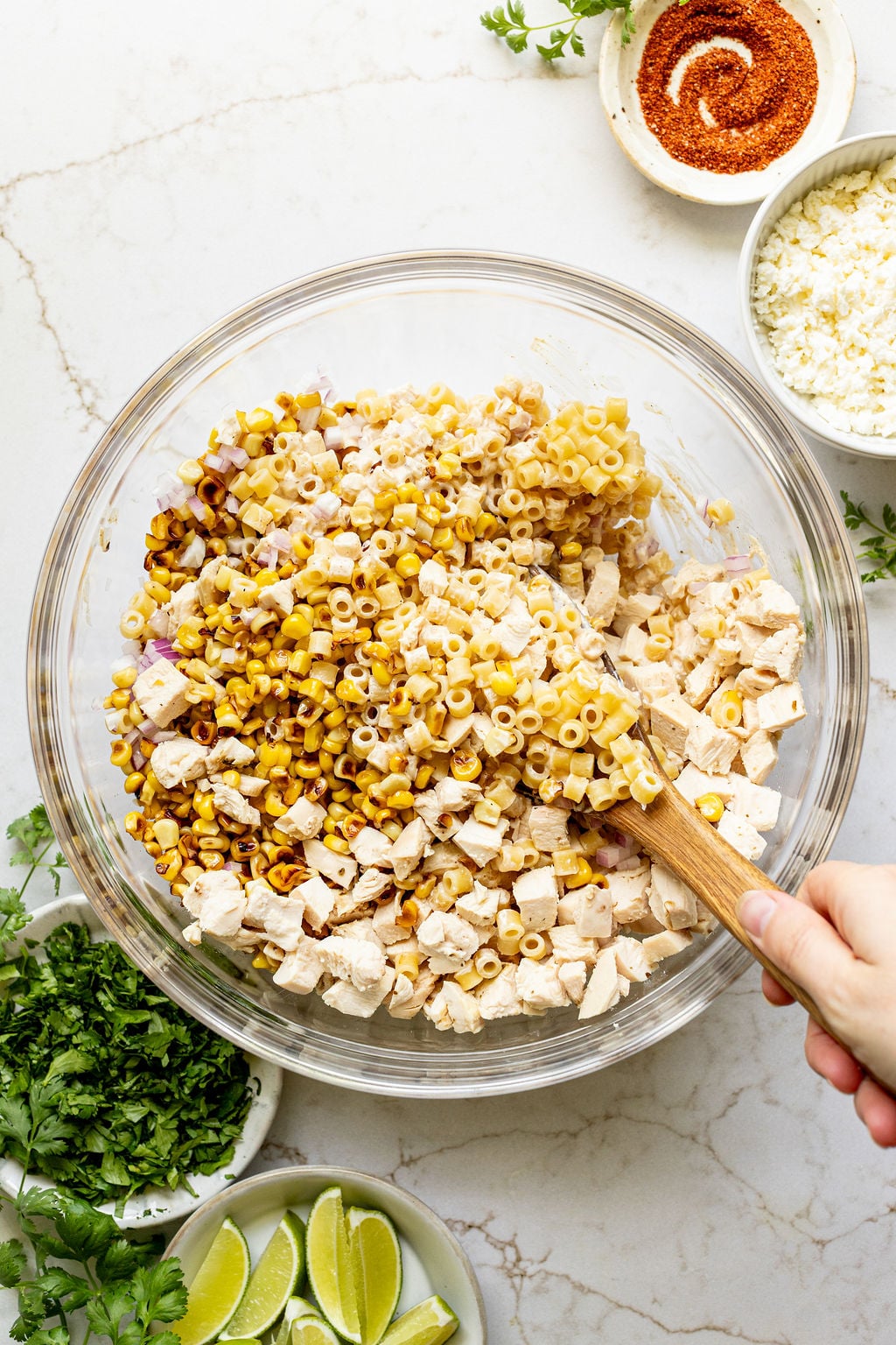 A hand stirring cooked pasta, diced chicken, red onion, and charred corn in a bowl.