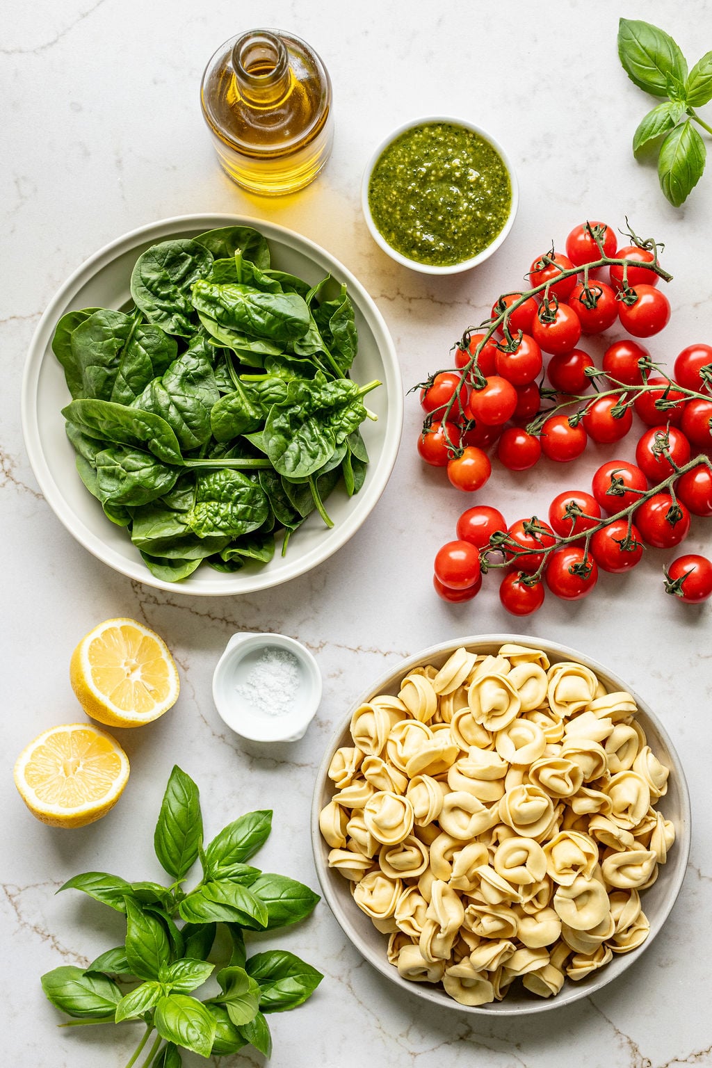 Bowls of tortellini, spinach, and pesto next to a lemon, basil, and cherry tomatoes.