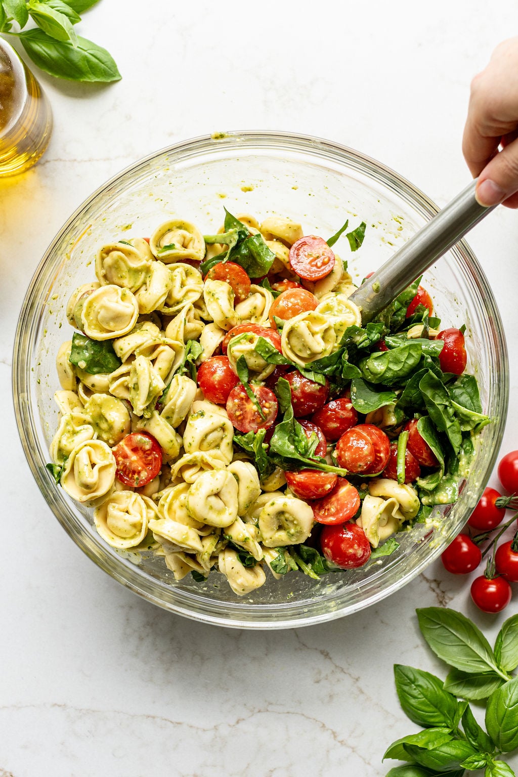 A hand stirring cooked tortellini, chopped spinach, and halved cherry tomatoes in a bowl.
