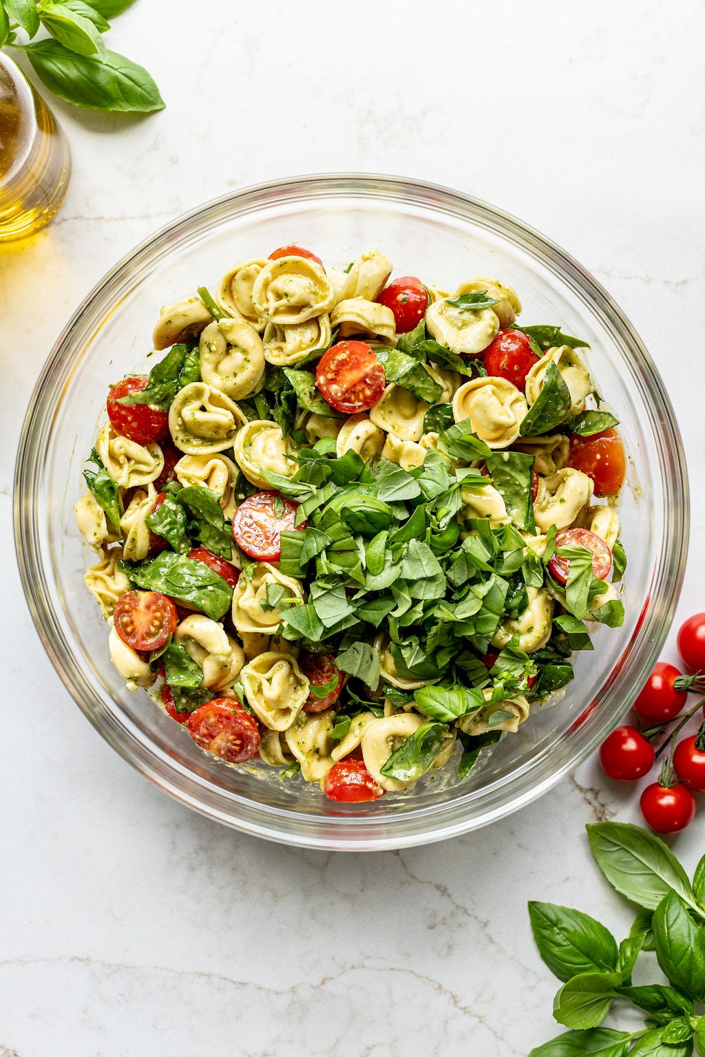 Cooked tortellini, cherry tomatoes, basil, spinach, and pesto in a glass bowl.