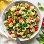 Fresh basil leaves on top of cooked tortellini, cherry tomatoes, fresh basil, spinach, and pesto in a bowl.