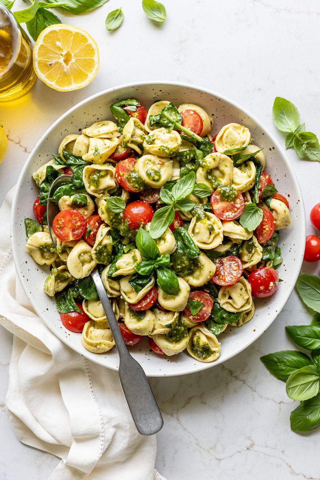 Fresh basil leaves on top of cooked tortellini, cherry tomatoes, fresh basil, spinach, and pesto in a bowl.