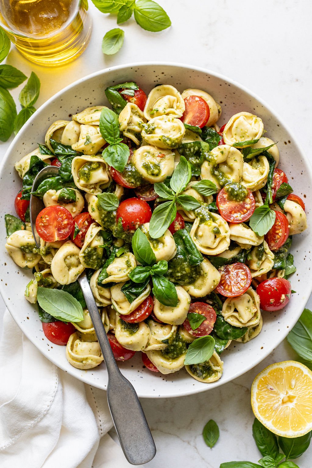 Cheese tortellini, cherry tomatoes, fresh basil, spinach, and pesto in a bowl next to a lemon slice.