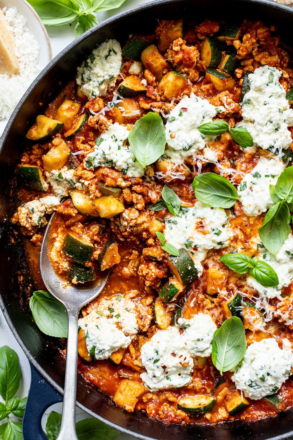 A spoon scooping saucy zucchini, ground turkey, red sauce, and ricotta from a skillet.