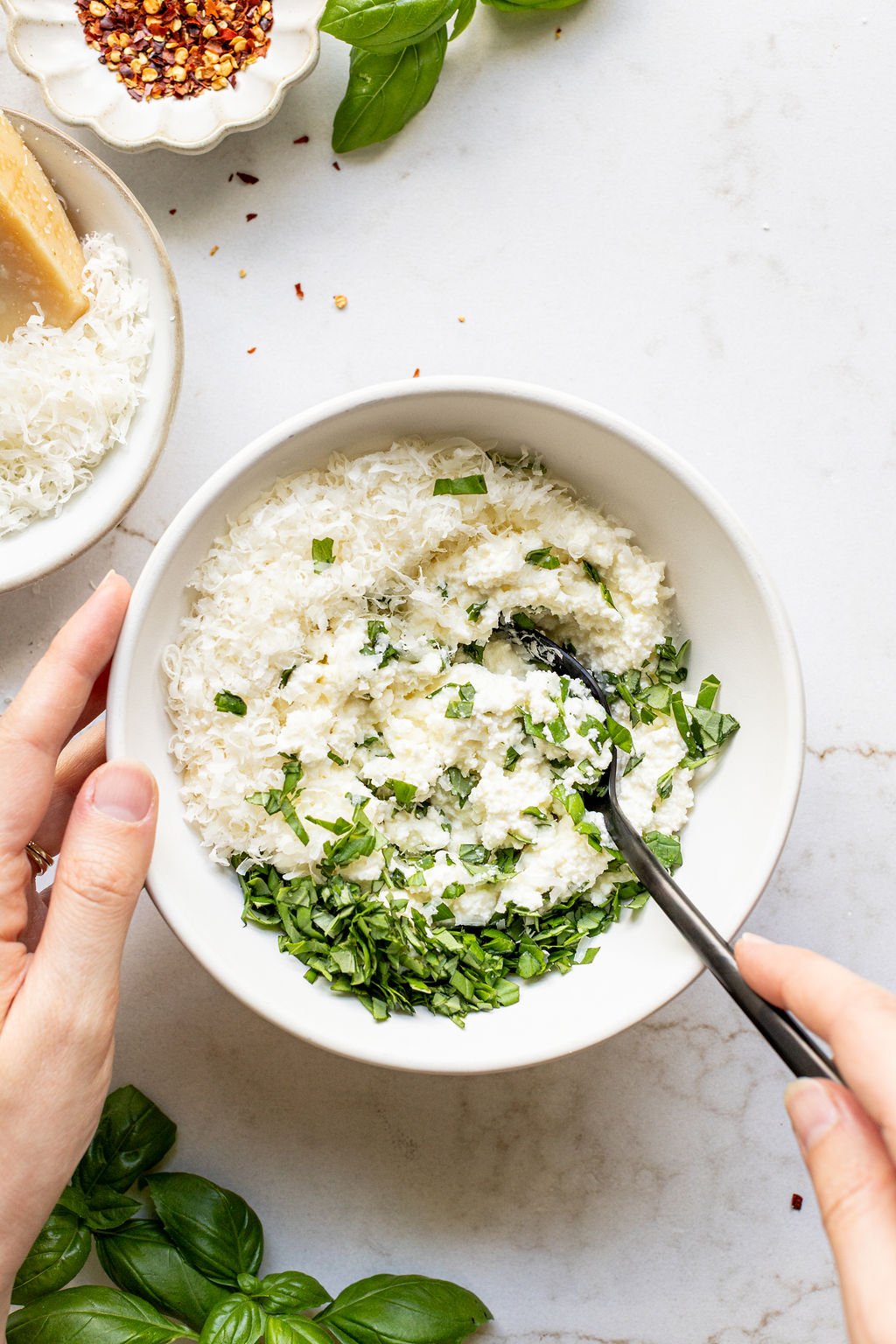 A hand stirring grated parmesan, ricotta, and chopped basil in a bowl.
