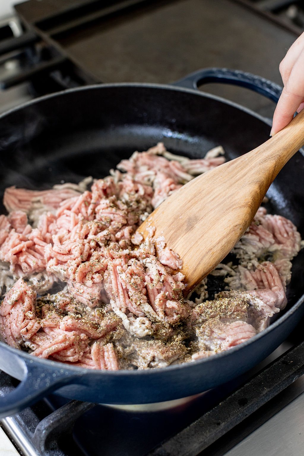 A hand breaking up ground turkey in a skillet with a wooden spoon.