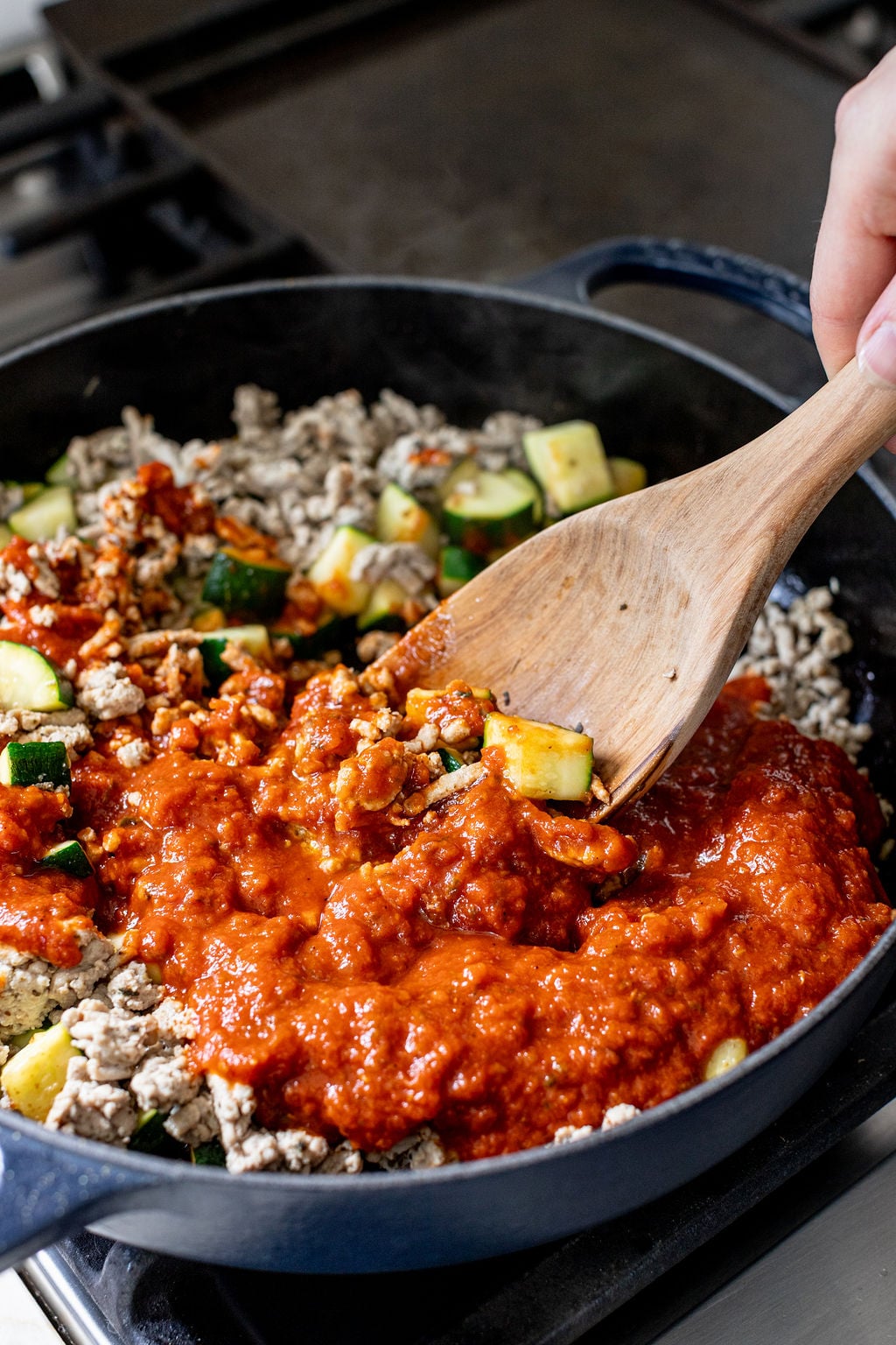 A hand stirring diced zucchini, ground turkey, and tomato sauce in a skillet.