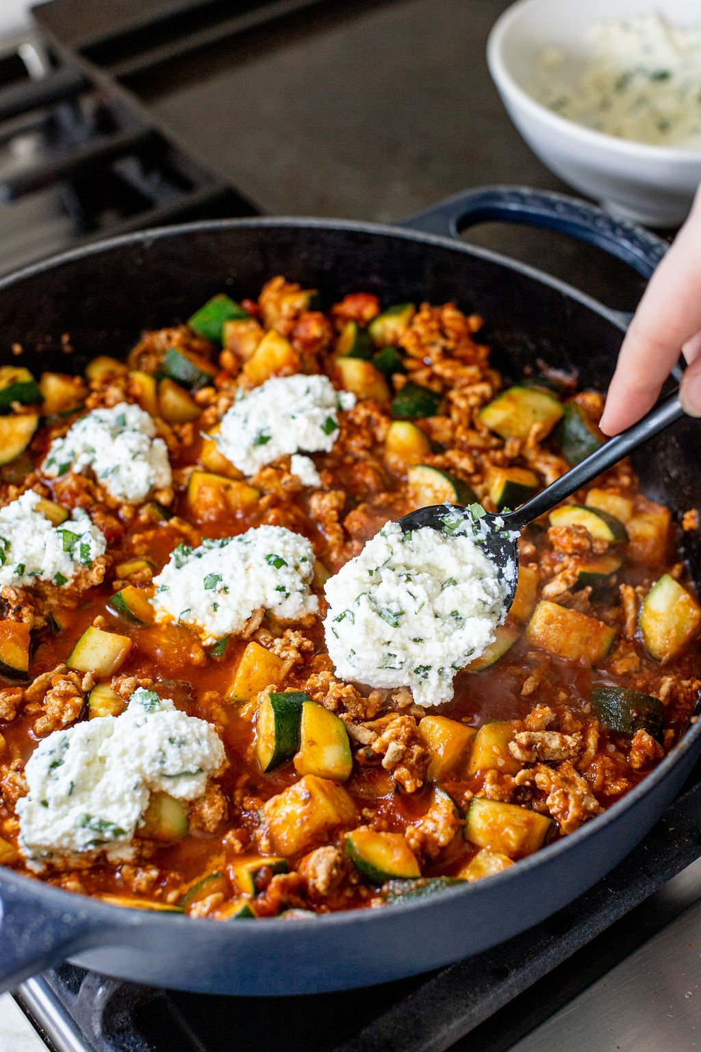 A spoon dolloping ricotta in a skillet of creamy red meat and veggies.