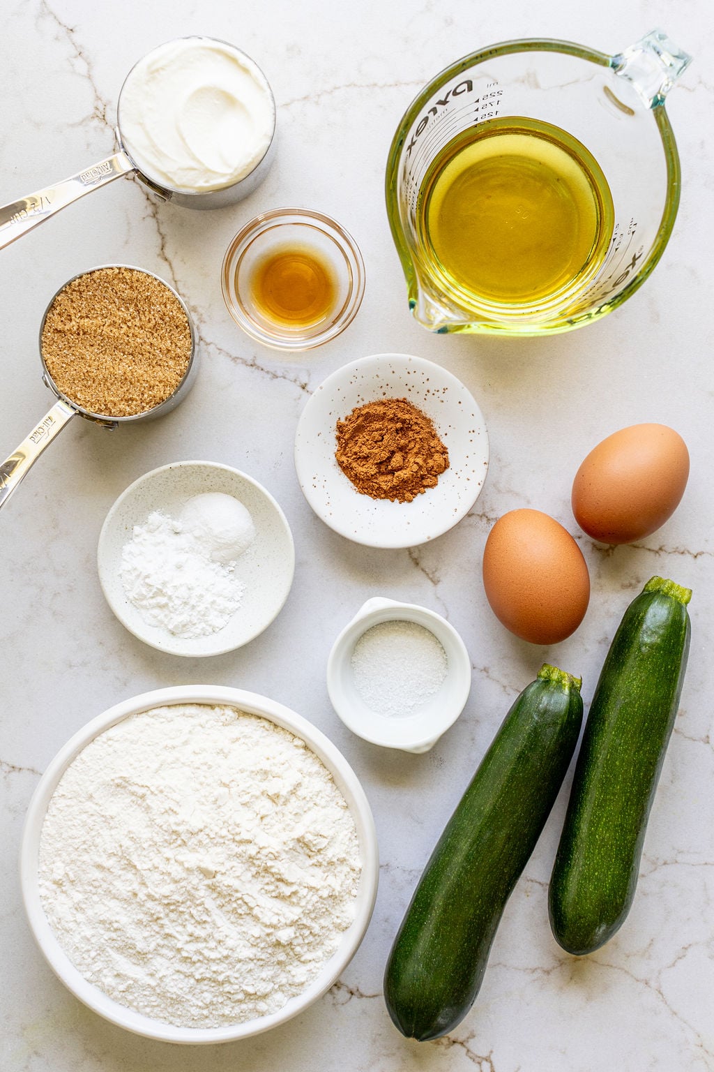 Bowls of flour, brown sugar, cinnamon, salt, vanilla, oil, and yogurt next to eggs and zucchini.