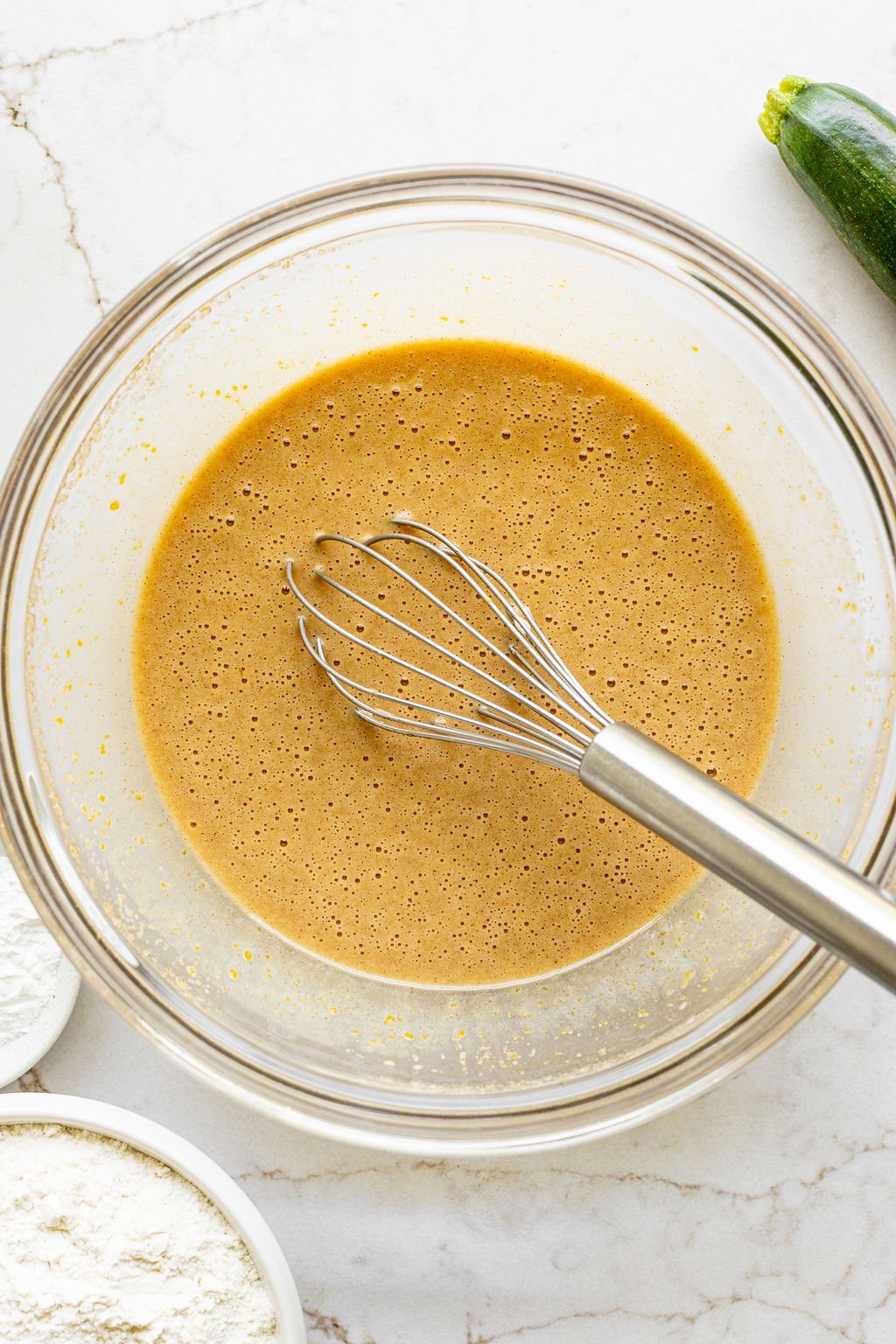A whisk sitting in a bowl of a golden brown, wet mixture.