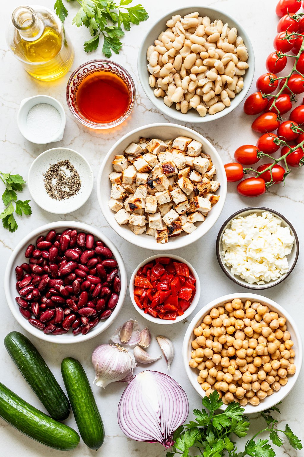 Top-down view of assorted salad ingredients including beans, grilled chicken, feta cheese, roasted red peppers, tomatoes, cucumbers, red onion, garlic, parsley, olive oil, vinegar, salt, and pepper.