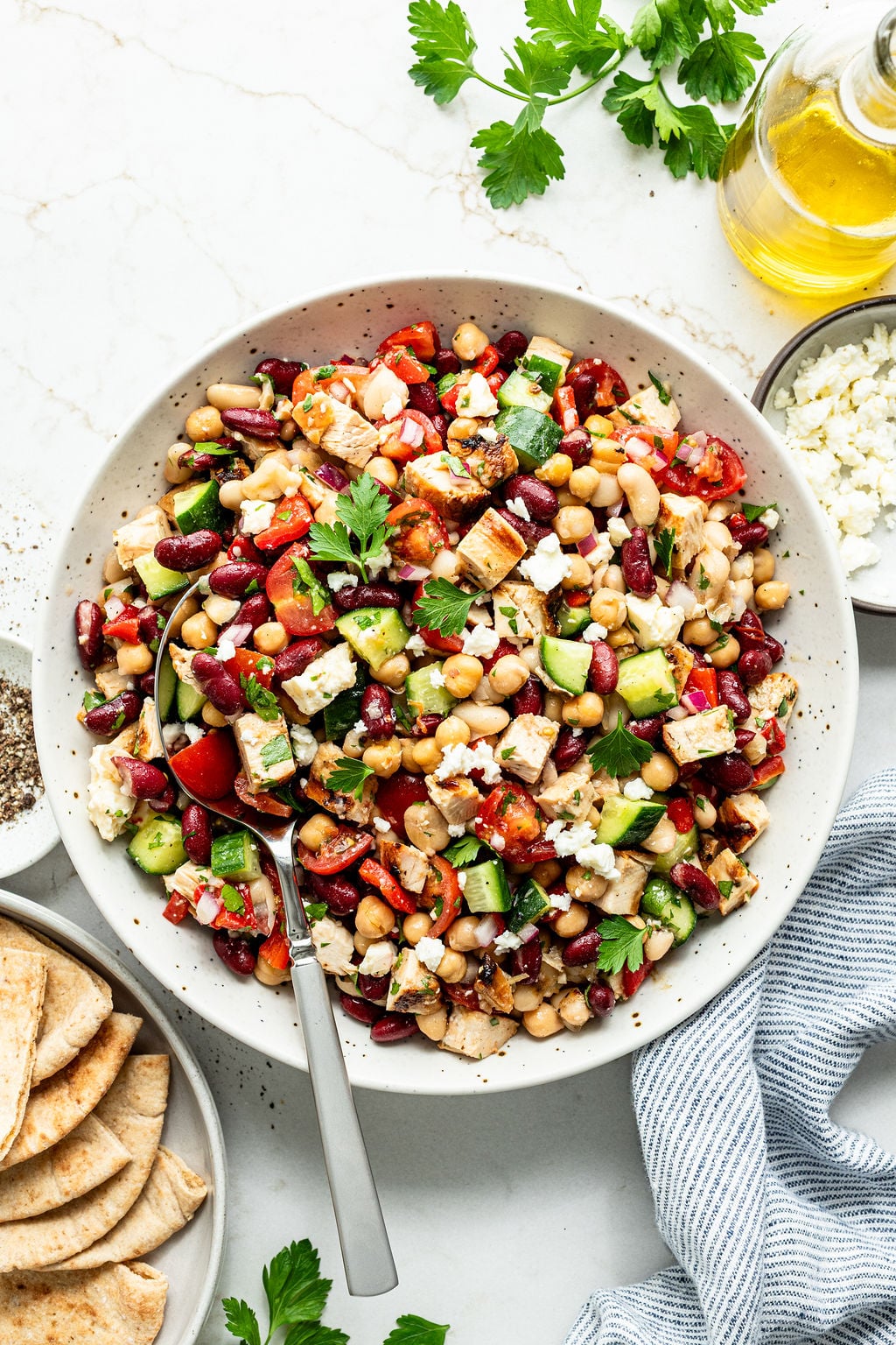 A bowl of mixed bean salad with cucumber, tomato, feta cheese, and parsley, served with pita bread and a bottle of olive oil.