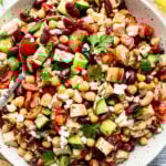 A bowl of mixed bean salad with chopped cucumbers, tomatoes, grilled chicken, feta cheese, parsley, and pita bread on the side.