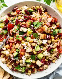 A bowl of mixed bean salad with chopped cucumbers, tomatoes, grilled chicken, feta cheese, parsley, and pita bread on the side.