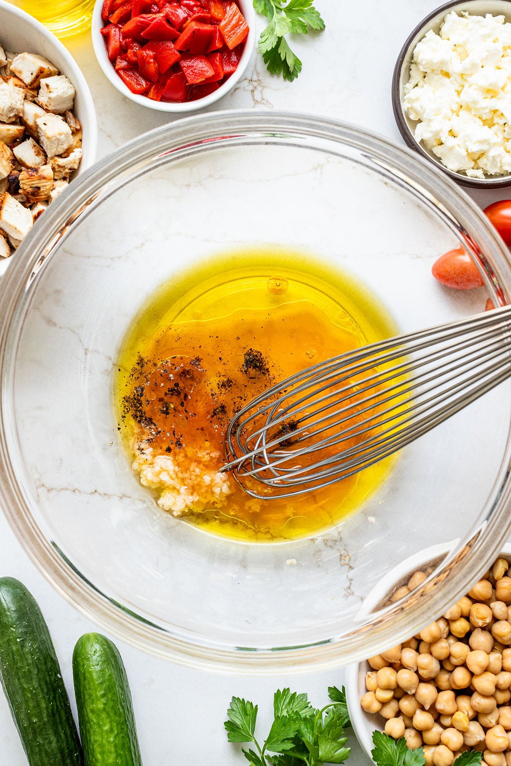 A whisk in a glass bowl containing oil, vinegar, minced garlic, salt, and pepper, surrounded by vegetables, chickpeas, cheese, and grilled chicken.