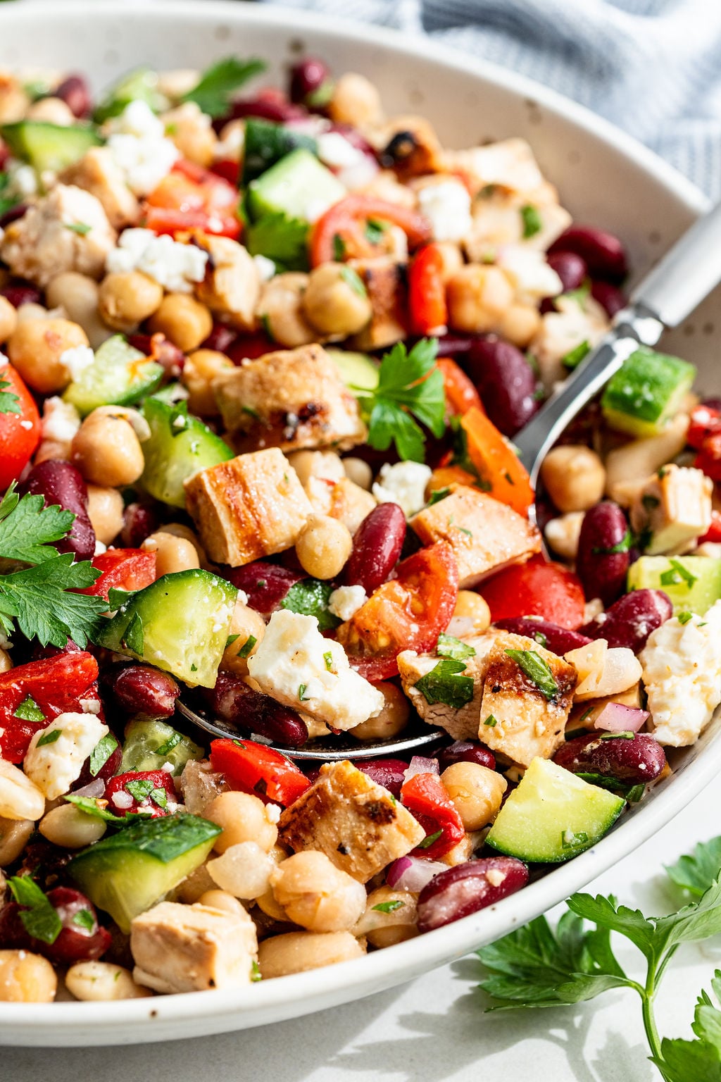 A close-up of a salad with chickpeas, kidney beans, grilled chicken, cucumber, tomato, feta cheese, red onion, and parsley in a white bowl with a fork.