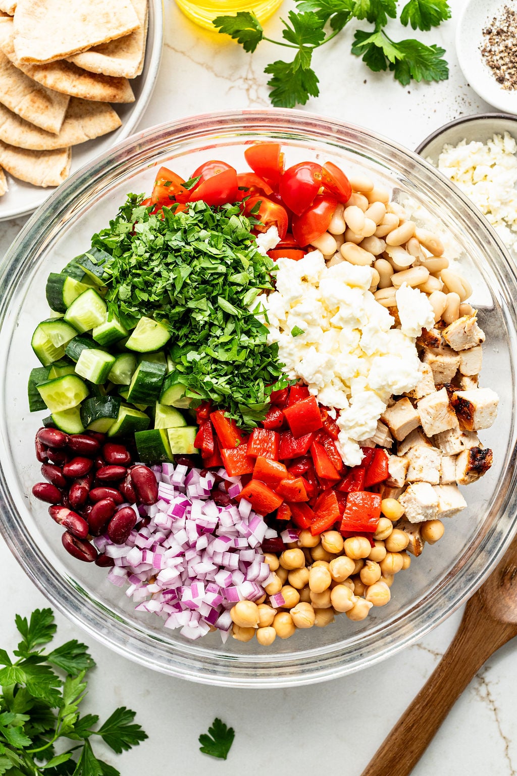 A glass bowl contains chopped cucumber, cherry tomatoes, grilled chicken, feta cheese, red onion, red bell pepper, chickpeas, white beans, kidney beans, and fresh parsley.