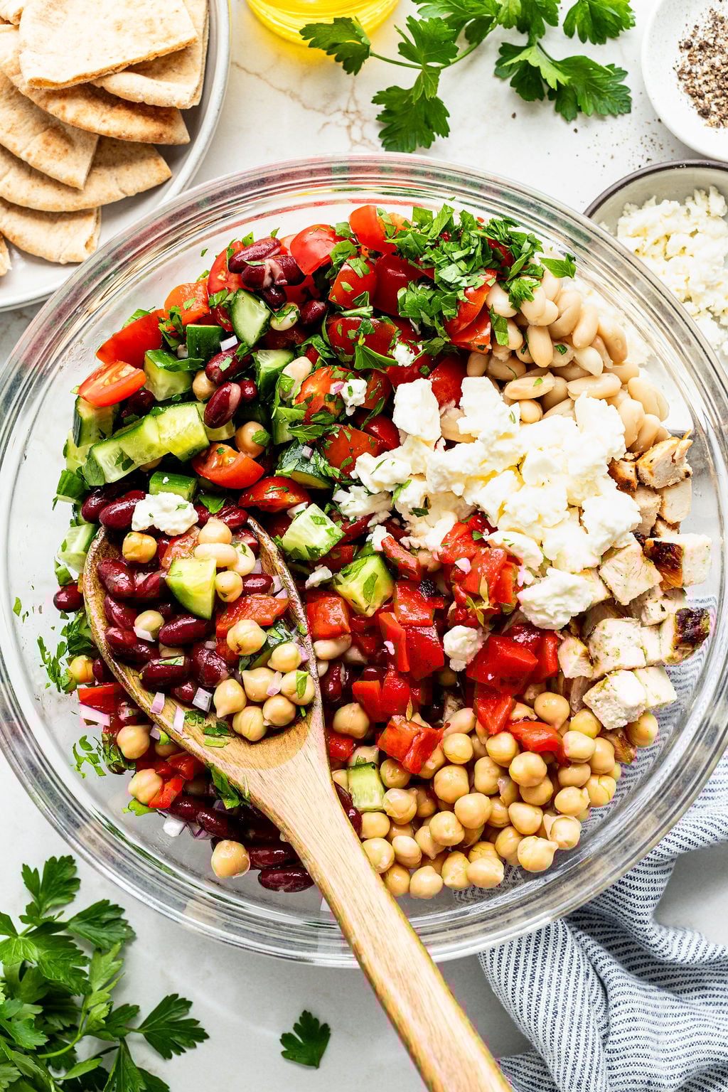 A clear glass bowl contains chopped tomatoes, cucumber, red onion, kidney beans, chickpeas, white beans, grilled chicken, parsley, and crumbled feta.