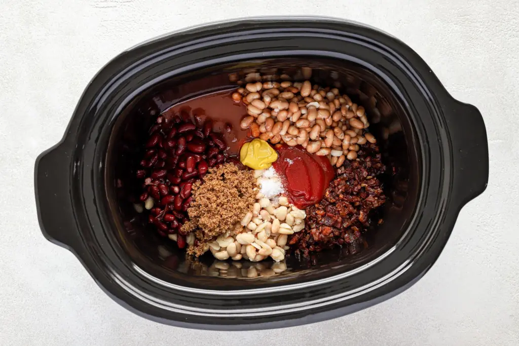 A slow cooker filled with kidney beans, pinto beans, white beans, brown sugar, tomato sauce, mustard, salt, and cooked ground beef, arranged in separate sections.