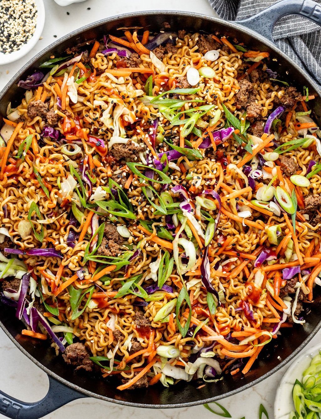 A skillet filled with cooked ramen noodles, ground beef, shredded carrots, cabbage, green onions, and sesame seeds.