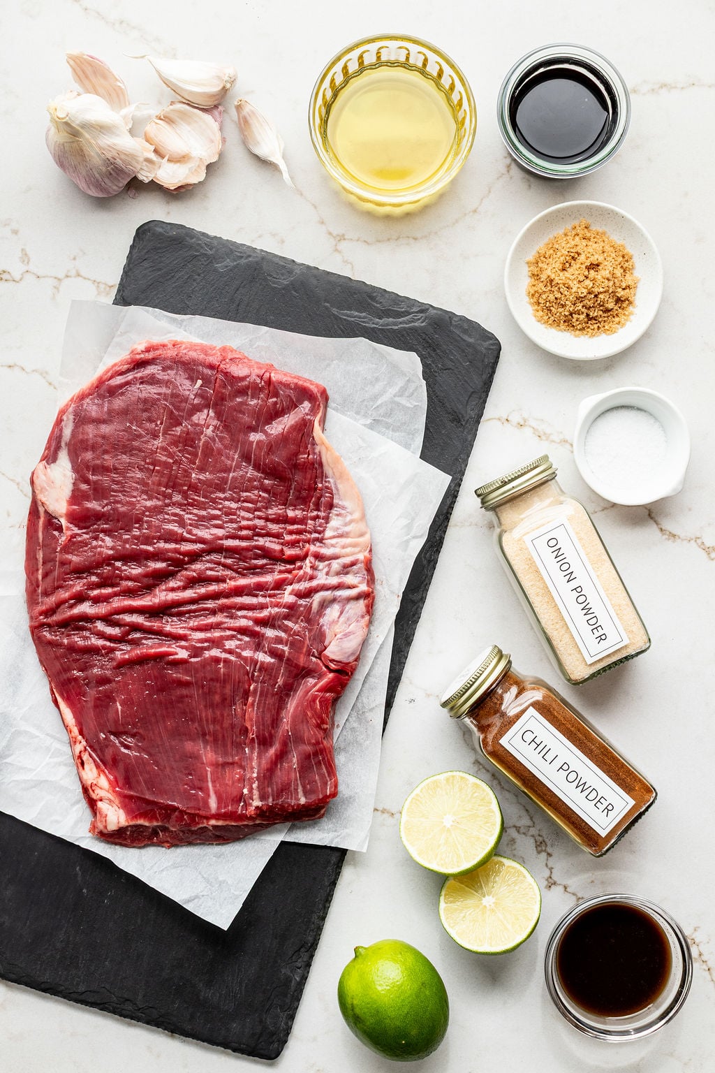 Raw steak, limes, spices, garlic cloves, and bowls of Worcestershire sauce, soy sauce, and oil on a table.