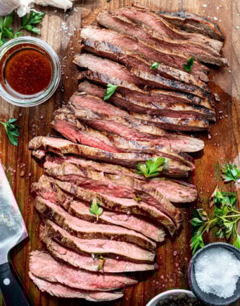 Slices of grilled flank steak on a wood cutting board next to dishes of salt and pepper.