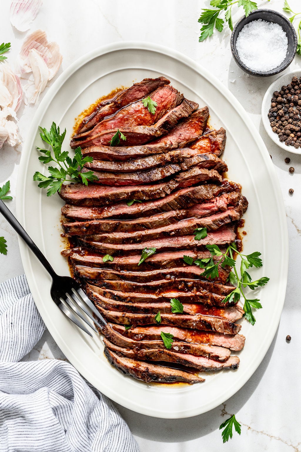 Slices of juicy, grilled flank steak on a platter topped with parsley.