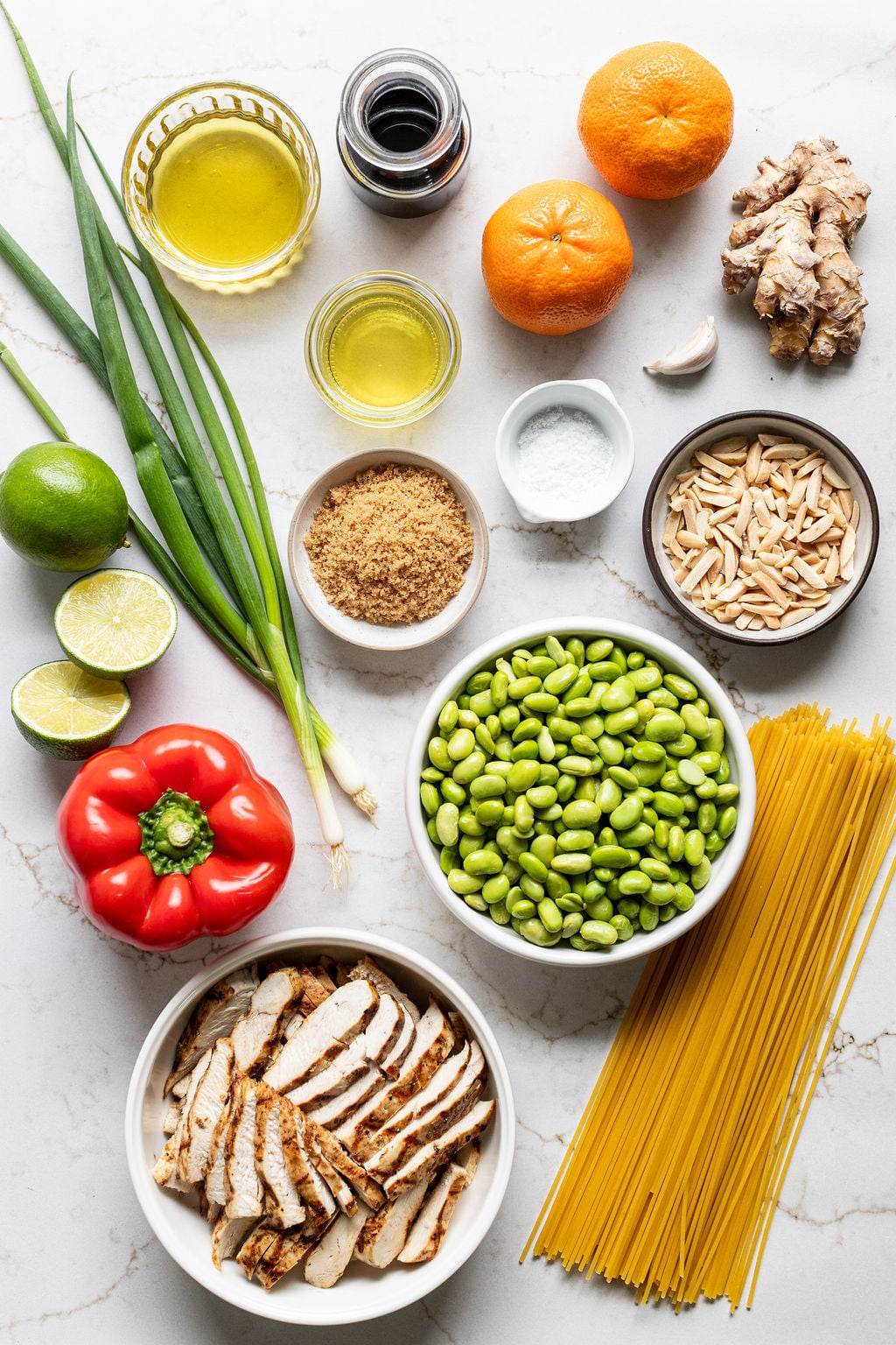 Assorted ingredients on a white surface, including grilled chicken, edamame, bell pepper, scallions, almonds, lime, mandarins, ginger, garlic, brown sugar, olive oil, soy sauce, and uncooked pasta.