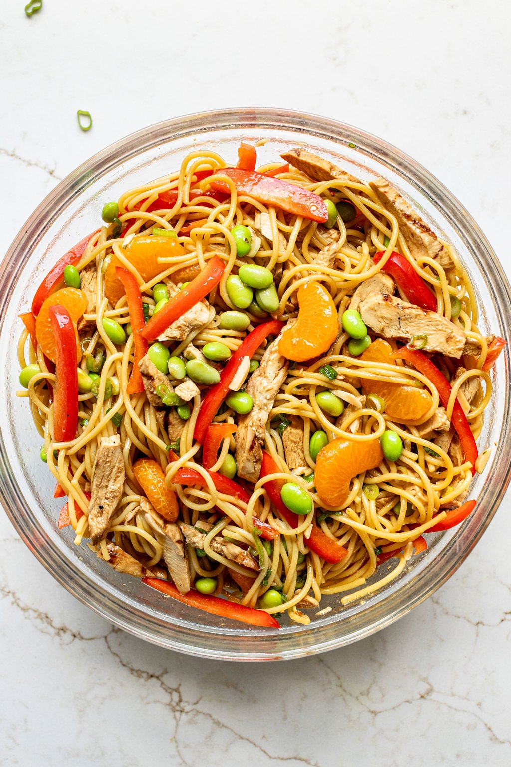 A bowl filled with pasta, chicken, sliced bell peppers, edamame, mandarins, chopped almonds, and green onions.