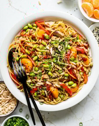 Bowls of mandarins, sesame seeds, almonds, and green onions next to a dish of sesame ginger noodles with veggies and chicken.