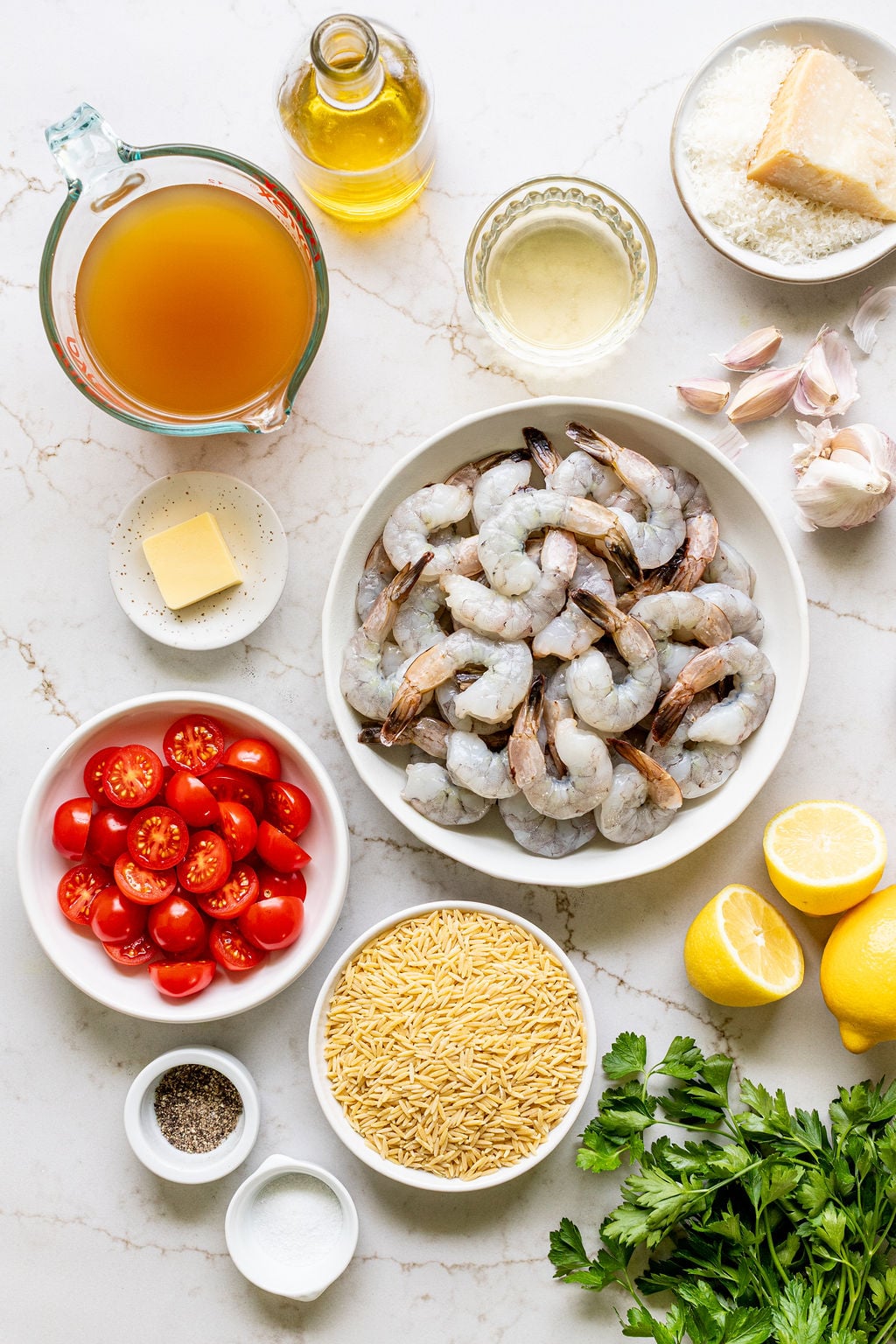 Overhead view of raw shrimp, orzo, cherry tomatoes, broth, olive oil, butter, garlic, parmesan, lemon, parsley, salt, and pepper arranged on a white countertop.