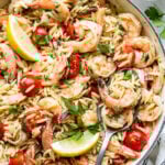 A skillet filled with orzo pasta, shrimp, cherry tomatoes, parsley, and lemon wedges, with a serving spoon resting inside.